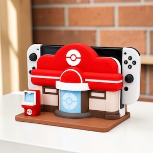 Peut inclure: Une station d'accueil pour console de jeu en forme de bâtiment rouge et blanc avec un emblème Poké Ball central. Le support présente une porte bleu clair, un petit kiosque rouge sur le côté et une base marron. Une console blanche est insérée.