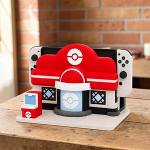 Op de afbeelding: Een rood-wit Nintendo Switch-dock ontworpen als een Pokémon Center-gebouw, met een centraal Poké Ball-embleem, ramen in glas-in-loodstijl en een kleine bijpassende rood-witte accessoirestandaard op een platte witte basis.