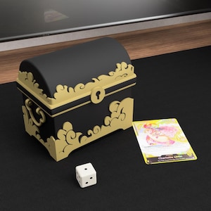 Könnte beinhalten: Eine schwarze und goldene Schatzkiste mit Schlüsselloch und Zierleisten, zusammen mit einem weißen Würfel mit zwei sichtbaren Punkten. Eine Sammelkarte mit dem Namen "Charlotte Linlin" ist ebenfalls vorhanden.