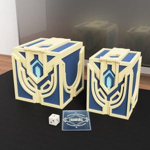 May include: Two blue and gold decorative boxes with a card and die. The boxes have a geometric design with a light blue accent. The card has the words "League Legends" on it. The die is white with black dots.