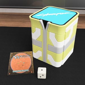 May include: A yellow and white rectangular card storage box with a light blue top featuring the text Cyberpunk, accompanied by a Magic The Gathering card and a small white six-sided die with black pips on a black surface.