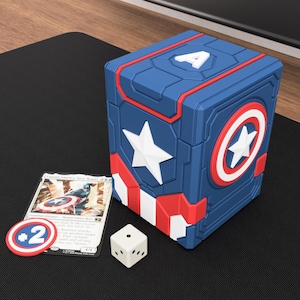 Può includere: Un portamazzo a tema Captain America blu, rosso e bianco con una stella bianca, un emblema a scudo, una lettera A bianca sulla parte superiore, un gettone +2 a forma di scudo, un dado a sei facce e una carta collezionabile.
