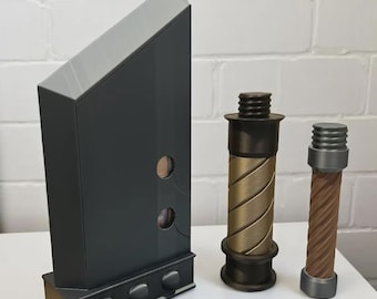 May include: A dark gray rectangular object with a light gray angled top, two cylindrical objects with threaded ends, one bronze and one brown, and a base with three circular accents. The objects are on a white surface.