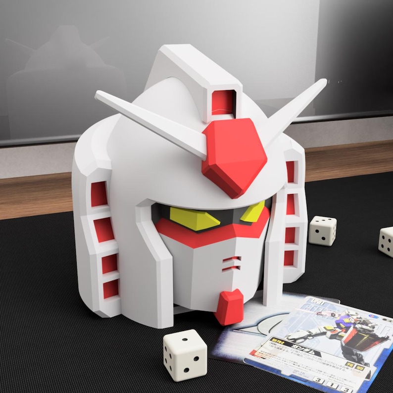 May include: A white and red Gundam head-shaped container with yellow accents. The container is on a black surface with white dice and trading cards. The container has a detailed design and is a collectible item.