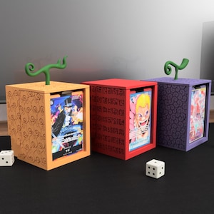 Könnte beinhalten: Drei farbenfrohe Kartenaufbewahrungsboxen, jede mit einem einzigartigen Design und einem grünen stielartigen Griff. Die Boxen sind orange, rot und lila, mit sichtbaren Kartenmotiven. Würfel sind vor zwei der Boxen platziert.