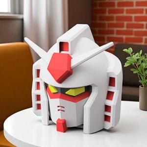 May include: A white and red Gundam robot head figurine with yellow accents. The figurine has a geometric design and is placed on a white table. The head features a red crest and details, with a stylized face.
