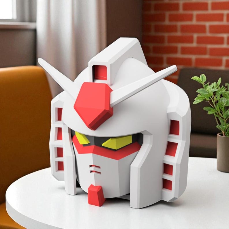 May include: A white and red Gundam robot head figurine with yellow accents. The figurine has a geometric design and is placed on a white table. The head features a red crest and details, with a stylized face.
