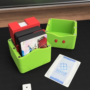 Könnte beinhalten: Eine limegrüne und rote Kunststoff-Kartenbox mit abnehmbarem Deckel, die ein Kartenspiel enthält. Die Box hat kleine kreisförmige Magnete an den Kanten. Eine einzelne Gundam Card Game-Karte liegt offen neben der Box auf einer schwarzen Fläche.