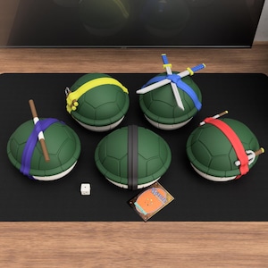 Op de afbeelding: Vijf groene schildpadvormige objecten met verschillende gekleurde hoofdbanden en wapens. Eén heeft een gele hoofdband, een andere blauw met zwaarden, een derde met een paarse hoofdband en een staf, een vierde met een rode hoofdband en sai, en de laatste met zwarte strepen. Een dobbelsteen en een kaart zijn ook aanwezig.