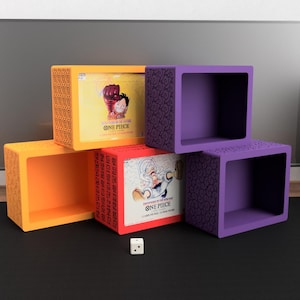 Könnte beinhalten: Eine Präsentation von farbenfrohen, offenen Würfelregalen in Orange, Gelb und Lila. Die Regale sind mit gemusterten Seiten verziert und enthalten Boxen mit "ONE PIECE"-Grafiken. Ein weißer Würfel mit schwarzen Punkten liegt davor.