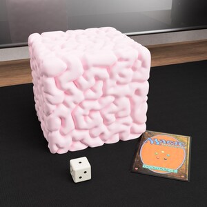 May include: A light pink, cube-shaped object with a textured, organic pattern. A white die with black dots and a Magic: The Gathering card are next to the cube. The items are on a black surface.