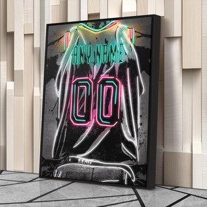 Op de afbeelding: Een ingelijste neonkunstprint van een basketbalshirt. Het shirt is zwart met de tekst "ANY NAME" in teal en het nummer "00" in roze en teal. Het kunstwerk heeft een neon gloed effect.