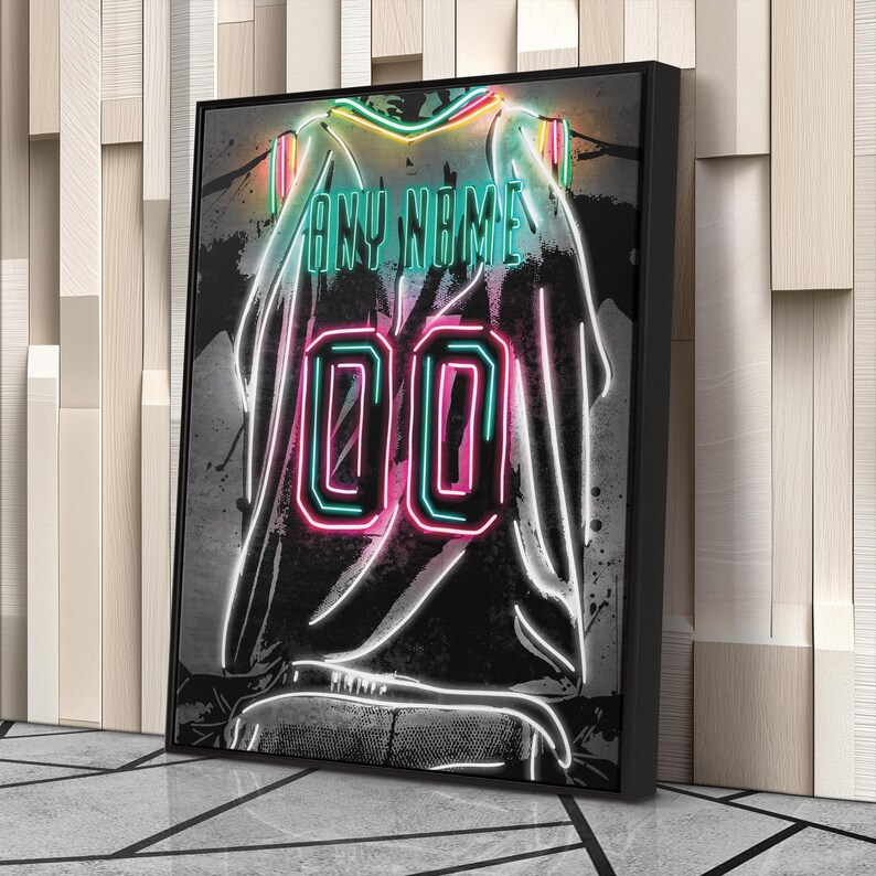 Op de afbeelding: Een ingelijste neonkunstprint van een basketbalshirt. Het shirt is zwart met de tekst "ANY NAME" in teal en het nummer "00" in roze en teal. Het kunstwerk heeft een neon gloed effect.