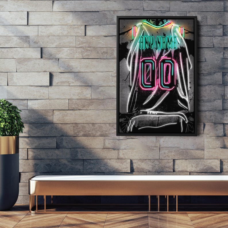 Op de afbeelding: Een ingelijst kunstwerk in neonstijl van een shirt met de tekst "ANY NAME" en het nummer "00". Het shirtontwerp heeft neonkleuren groen, roze en wit tegen een donkere achtergrond. Het kunstwerk wordt tentoongesteld op een bakstenen muur.