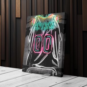 Op de afbeelding: Canvas print van een neon-stijl jersey met de tekst "ANY NAME" boven het nummer "00". Het jersey-ontwerp heeft een zwarte achtergrond met witte contouren en spetters van kleuren, waaronder groen en roze.