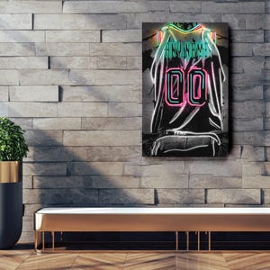 Op de afbeelding: Een neon-stijl kunstwerk van een basketbalshirt met de tekst "ANY NAME" en het nummer "00". Het shirtontwerp heeft heldere neon groene, roze en witte contouren tegen een donkere achtergrond. Het kunstwerk is op een bakstenen muur gemonteerd.