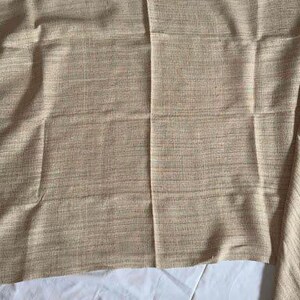 May include: A tan-colored fabric with a woven texture. The fabric is folded and lies on a white surface. The fabric has a subtle pattern of light and dark threads, creating a textured appearance.