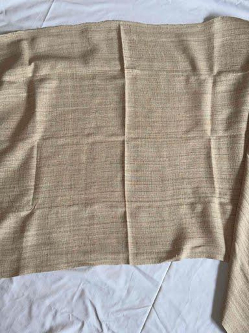 May include: A tan-colored fabric with a woven texture. The fabric is folded and lies on a white surface. The fabric has a subtle pattern of light and dark threads, creating a textured appearance.