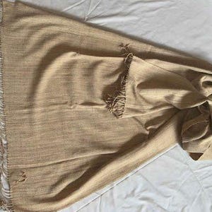 May include: A tan-colored woven scarf with fringed edges. The scarf is folded and draped, showcasing its texture and design. The fringe adds a decorative element to the scarf.