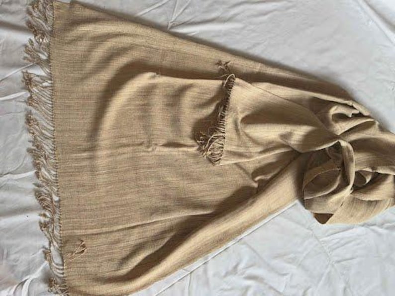 May include: A tan-colored woven scarf with fringed edges. The scarf is folded and draped, showcasing its texture and design. The fringe adds a decorative element to the scarf.