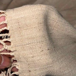 May include: Close-up of a beige woven fabric with a textured surface. The edge features a decorative, openwork crochet trim. The fabric appears to be soft and pliable, with a natural, organic aesthetic.