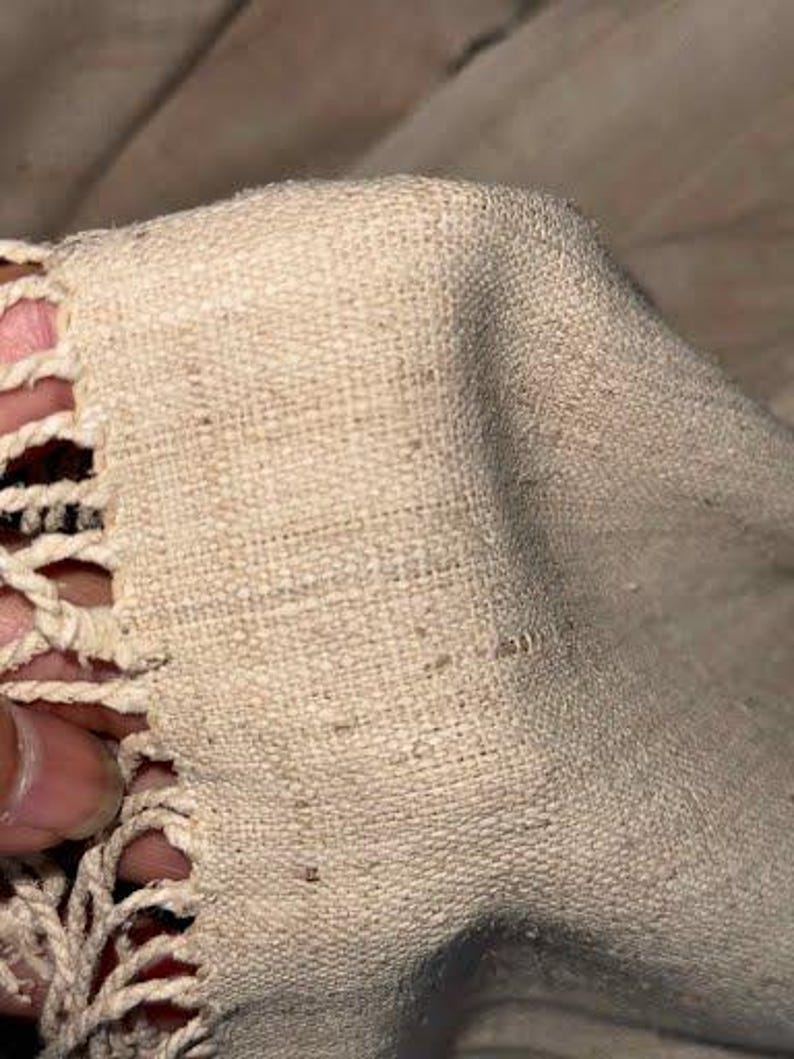 May include: Close-up of a beige woven fabric with a textured surface. The edge features a decorative, openwork crochet trim. The fabric appears to be soft and pliable, with a natural, organic aesthetic.