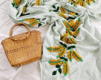 May include: A light blue scarf with embroidered yellow and green floral design, next to a woven wicker handbag with a bamboo handle. The scarf is draped on a white surface.