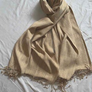 May include: A beige scarf with a fringed edge is draped on a white surface. The scarf is made of a woven fabric and has a soft, flowing appearance. The scarf is folded and draped to show its texture and design.