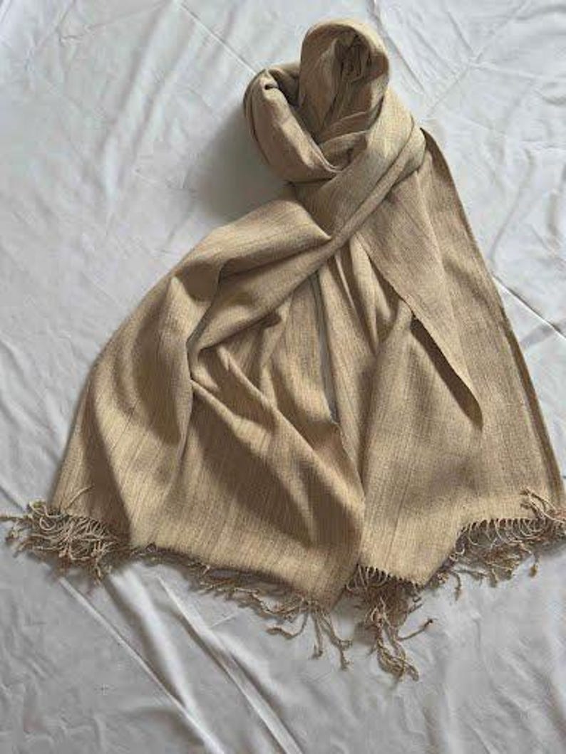 May include: A beige scarf with a fringed edge is draped on a white surface. The scarf is made of a woven fabric and has a soft, flowing appearance. The scarf is folded and draped to show its texture and design.