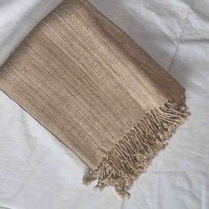 May include: Folded tan-colored woven scarf with fringe detail. The scarf is made of a textured material and is folded neatly. The fringe is at the bottom edge of the scarf. The scarf is resting on a white surface.
