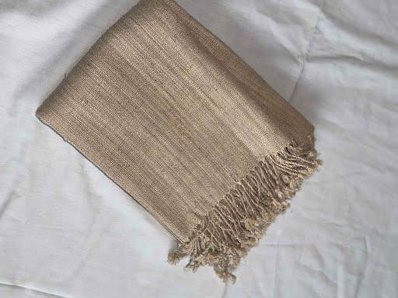 May include: Folded tan-colored woven scarf with fringe detail. The scarf is made of a textured material and is folded neatly. The fringe is at the bottom edge of the scarf. The scarf is resting on a white surface.