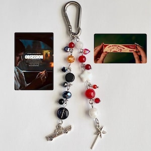 May include: A keyring featuring a silver carabiner with two dangling strands of beads in red, white, black, and orange. One strand ends with a silver car charm, and the other ends with a silver star wand charm. Two small rectangular photo inserts are included.