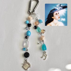 May include: A keychain featuring a silver carabiner attached to two strands of beads in shades of blue, black, peach, and clear. The strands include charms shaped like a television and a musical note, alongside a small square image of a person.