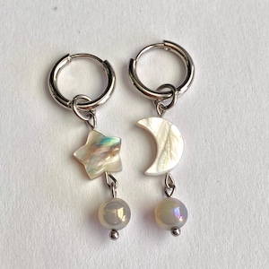 May include: A pair of silver hoop earrings. One earring features a star-shaped charm, the other a crescent moon. Both have iridescent shell accents and a dangling iridescent bead. The earrings are designed for pierced ears.
