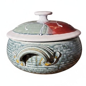 May include: A round ceramic lidded casserole dish featuring a textured, mottled blue-gray glaze with gold-toned accents. The lid displays a segmented design with sections of muted green, deep red, and white, finished with a central knob handle.