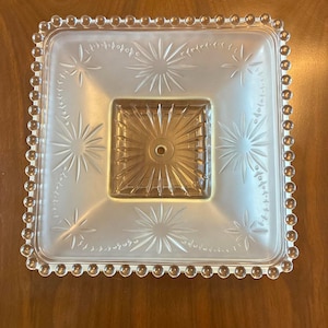 May include: A square glass light fixture cover featuring a frosted finish with clear starburst patterns. The center has a clear, raised square section with radiating lines and a central hole. The outer edge is bordered by a row of clear glass beads.