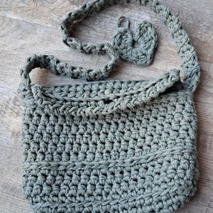 Op de afbeelding: Een handgemaakte gehaakte schoudertas in saliegroen. De tas heeft een dik, getextureerd steekpatroon en een gevlochten band. Een klein, hartvormig detail is aan de band bevestigd. De tas is ontworpen met een afgeronde bodem en een klepsluiting.