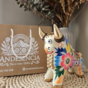 May include: A ceramic bull figurine with gold accents and colorful floral designs. The bull has a cream-colored body, gold horns, and is decorated with blue, pink, and yellow flowers. A brown paper shopping bag with the text "ANDESENCIA Peruvian Gifts" is in the background.