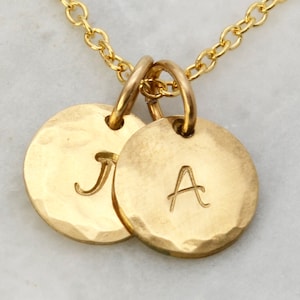 May include: Gold-colored necklace with two circular pendants. Each pendant has a hammered texture and a single initial engraved on it. The chain and connecting rings are also gold.
