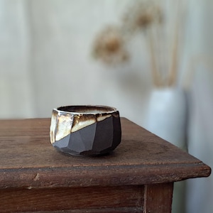 Peut inclure: Un petit bol en céramique fait à la main avec un design géométrique. Le bol présente un extérieur brun foncé avec une surface texturée et un bord intérieur plus clair, de couleur crème. Le bol est présenté sur une surface en bois.