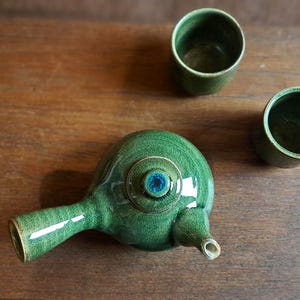 Può includere: Una teiera in ceramica verde fatta a mano con un centro blu e due tazze abbinate. La teiera ha un lungo beccuccio e un corpo arrotondato. Le tazze e la teiera sono su una superficie di legno.