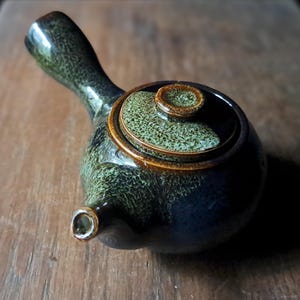May include: A ceramic teapot with a dark green and brown speckled glaze. The teapot has a handle, a spout, and a lid with a small knob. The teapot is on a wooden surface.