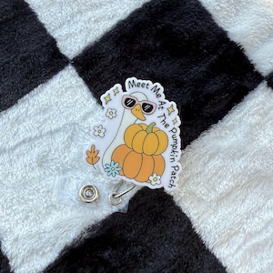 May include: A white and orange badge reel featuring a cartoon goose wearing sunglasses and sitting on pumpkins. The text "Meet Me At The Pumpkin Patch" is written around the goose. The badge reel has a clear plastic strap and a metal clip.