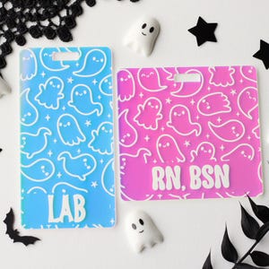 May include: Two rectangular acrylic badge reels featuring a repeating white ghost and star pattern. The left badge is blue with the white text LAB at the bottom. The right badge is pink with the white text RN, BSN at the bottom. Both have a slot at the top.