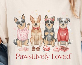 Puede incluir: Camiseta color crema con una ilustración en acuarela de cuatro perros, cada uno adornado con accesorios como una pajarita y un suéter. La frase "Pawsitively Loved" está impresa en escritura roja debajo. La camiseta es de estilo casual y cómodo.