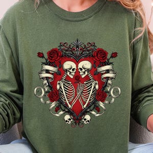 Puede incluir: Sudadera verde de manga larga con un diseño en forma de corazón con dos esqueletos tomados de la mano. El diseño incluye rosas rojas, adornos decorativos y una pancarta. Los esqueletos son blancos y el corazón es rojo.