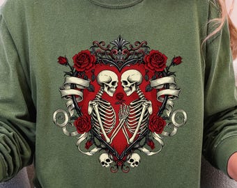 May include: Green long-sleeve sweatshirt featuring a heart-shaped design with two skeletons holding hands. The design includes red roses, decorative scrollwork, and a banner. The skeletons are white, and the heart is red.