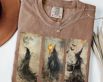 May include: Brown Comfort Colors t-shirt with a graphic of three witch scenes. The scenes depict witches with a full moon, a lantern, and a cauldron. A silver necklace with a pendant is on the shirt.