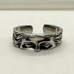 May include: A silver-colored adjustable ring featuring a sculpted face design. The ring has an open design and is detailed with wavy hair and facial features. The ring is likely made of a metal alloy.
