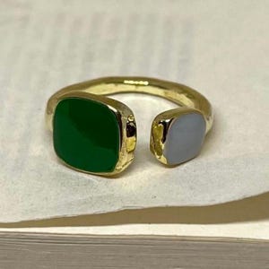 Könnte beinhalten: Ein goldfarbener Ring mit offenem Design. Ein Ende hat einen quadratischen, smaragdgrünen Stein, während das andere einen quadratischen, hellblauen Stein hat. Der Ring liegt auf einer weißen Oberfläche.